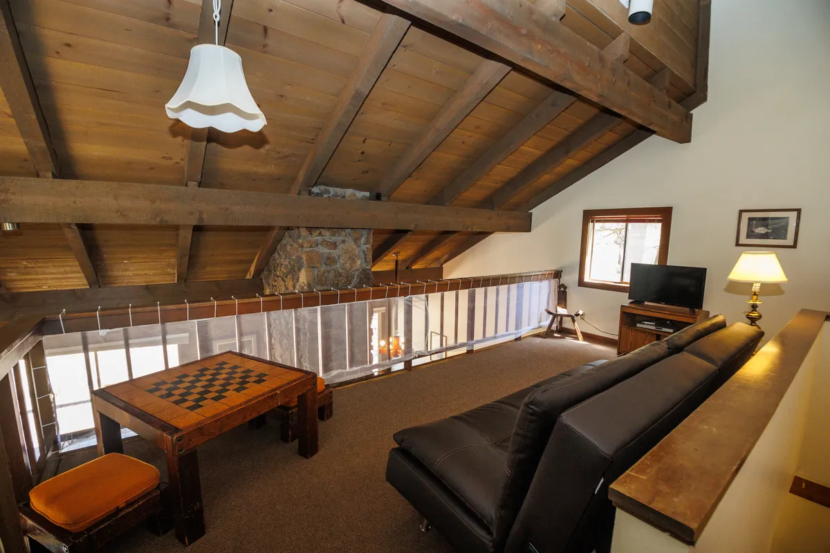 This image features a cozy loft area with exposed wooden beams, a chess table, a sofa, a lamp, and a television. The arrangement exudes a relaxed atmosphere, ideal for casual lounging, playing games, or movie nights.