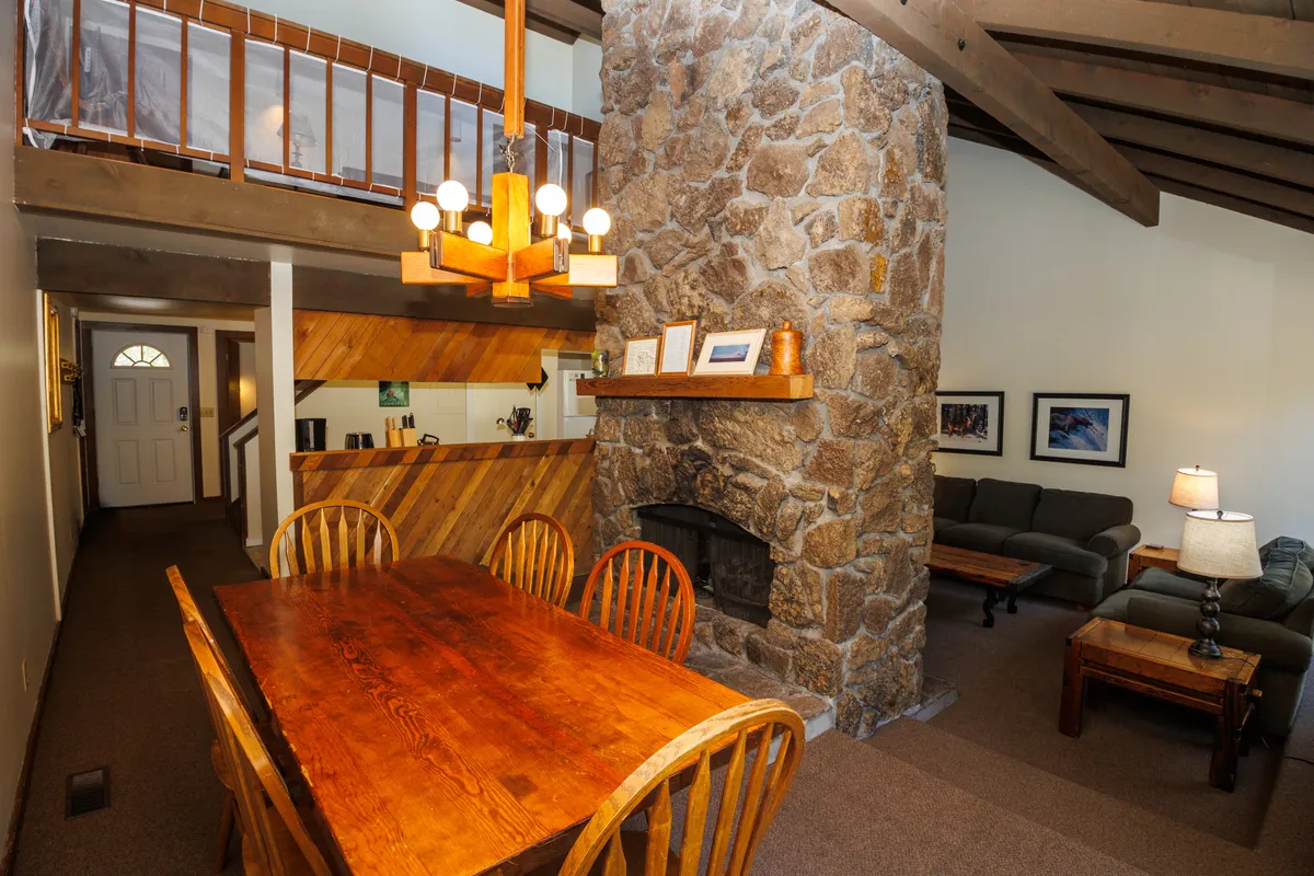 A bright, welcoming dining area showcases a long table set for a group, adjacent to a living area with a plush sofa and a prominent stone fireplace. The scene evokes togetherness and warmth, ideal for gathering after a day spent outdoors.
