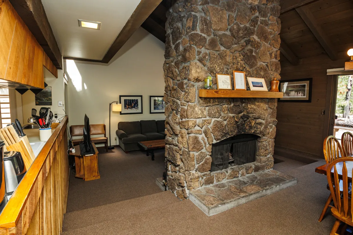 This image captures a warm and inviting open-concept space, featuring a stone-faced fireplace, a wooden dining table, a cozy seating area, and a kitchen counter. The fireplace is a central architectural element, dividing the living and dining areas, while large windows (implied by the lighting and layout) suggest a connection to the outdoors.