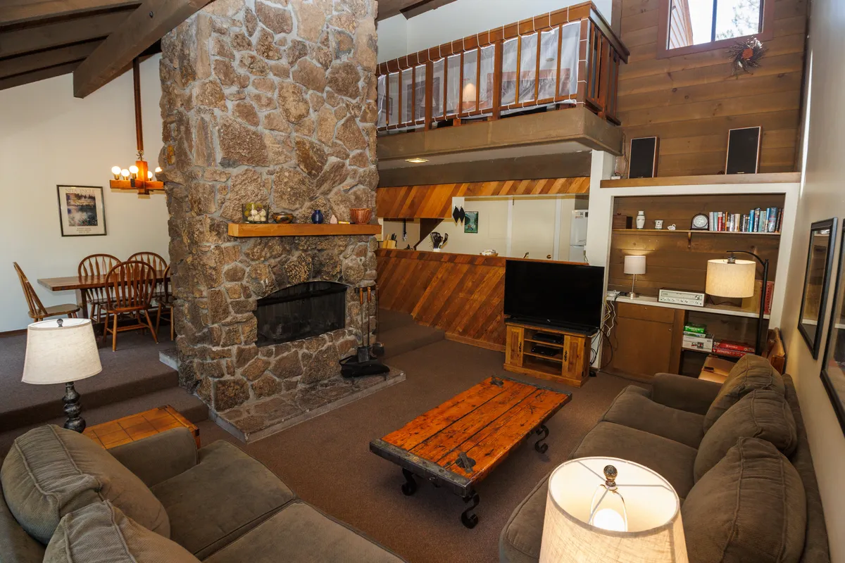 The image showcases a welcoming living area with a rustic stone, double-sided fireplace as the centerpiece, flanked by a warm wooden coffee table and ample seating. The open loft area offers additional space to relax, while a cozy dining table invites guests to gather for meals. The overall scene is bathed in natural light, creating an inviting, cabin-like atmosphere perfect for unwinding after outdoor adventures.