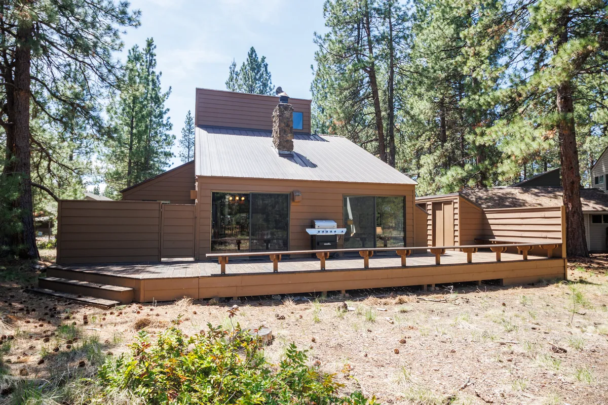 This exterior image showcases a spacious wooden deck attached to the cabin, with a prominent stone chimney and large windows that hint at the cozy interior. The deck is surrounded by trees, offering both privacy and a close connection to nature, making it ideal for outdoor dining and relaxation after a day of adventure.