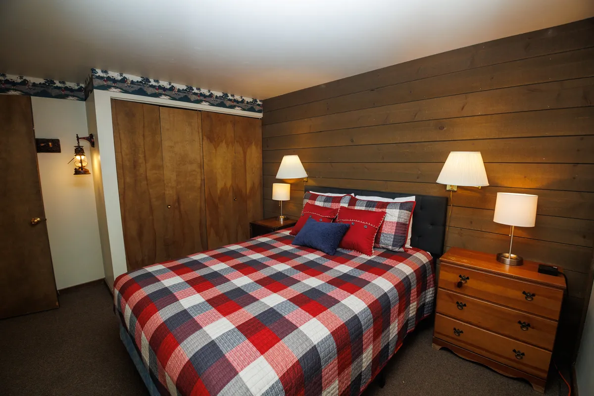 A cozy bedroom with a bed featuring plaid bedding, nightstands with lamps on either side, a wooden accent wall, and closet doors. Soft lighting and warm tones create a soothing environment ideal for unwinding.