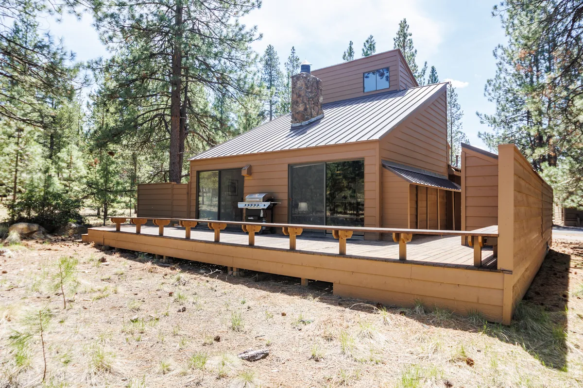 This exterior image features a spacious wooden deck connected to the home, framed by large windows and a stone chimney, and surrounded by tall trees. The setup clearly signals an inviting outdoor living space, perfect for barbecuing, dining al fresco, or relaxing amid nature.