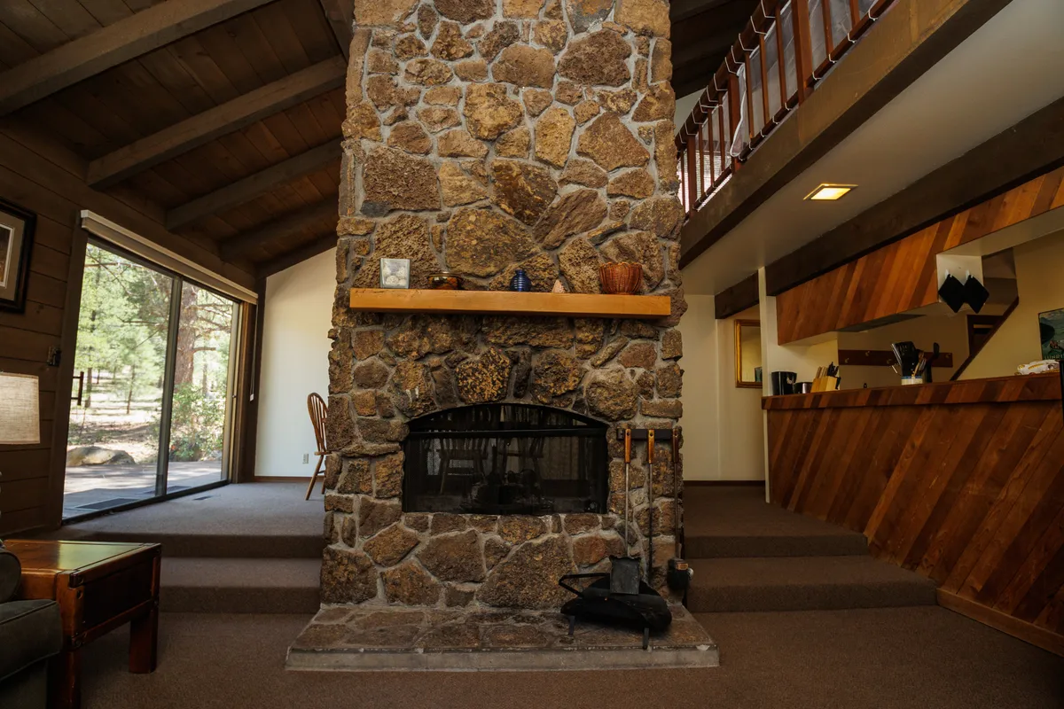 A spacious open-concept living area featuring a striking stone fireplace with a wooden mantel, cozy seating, and large windows that flood the room with natural light. The open layout invites gatherings and offers a clear view from the living space into the dining area.