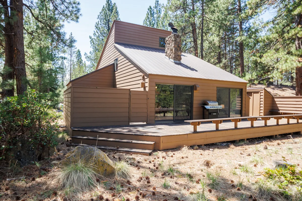 This exterior angle features a large wooden deck equipped for outdoor seating, a striking stone chimney, and expansive glass doors opening from the living areas. The mature trees in the background emphasize privacy and the home’s integration with its natural surroundings—a perfect spot for family gatherings or quiet mornings before a day of activities.