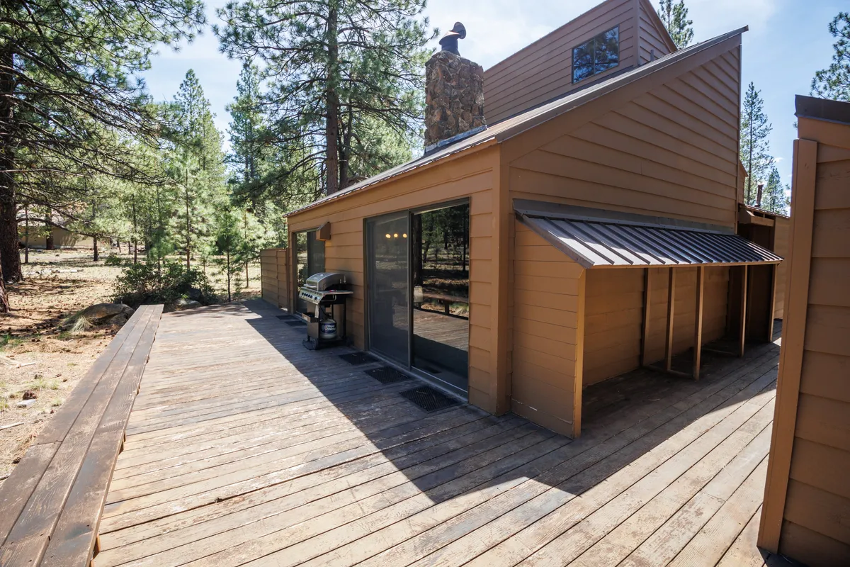 A welcoming exterior deck scene showcasing a barbecue grill, generous seating, large windows, and a chimney, all surrounded by mature trees. The photo captures the patio as a lively, functional space for gathering, grilling, and transitioning to Sunriver’s outdoor experiences.