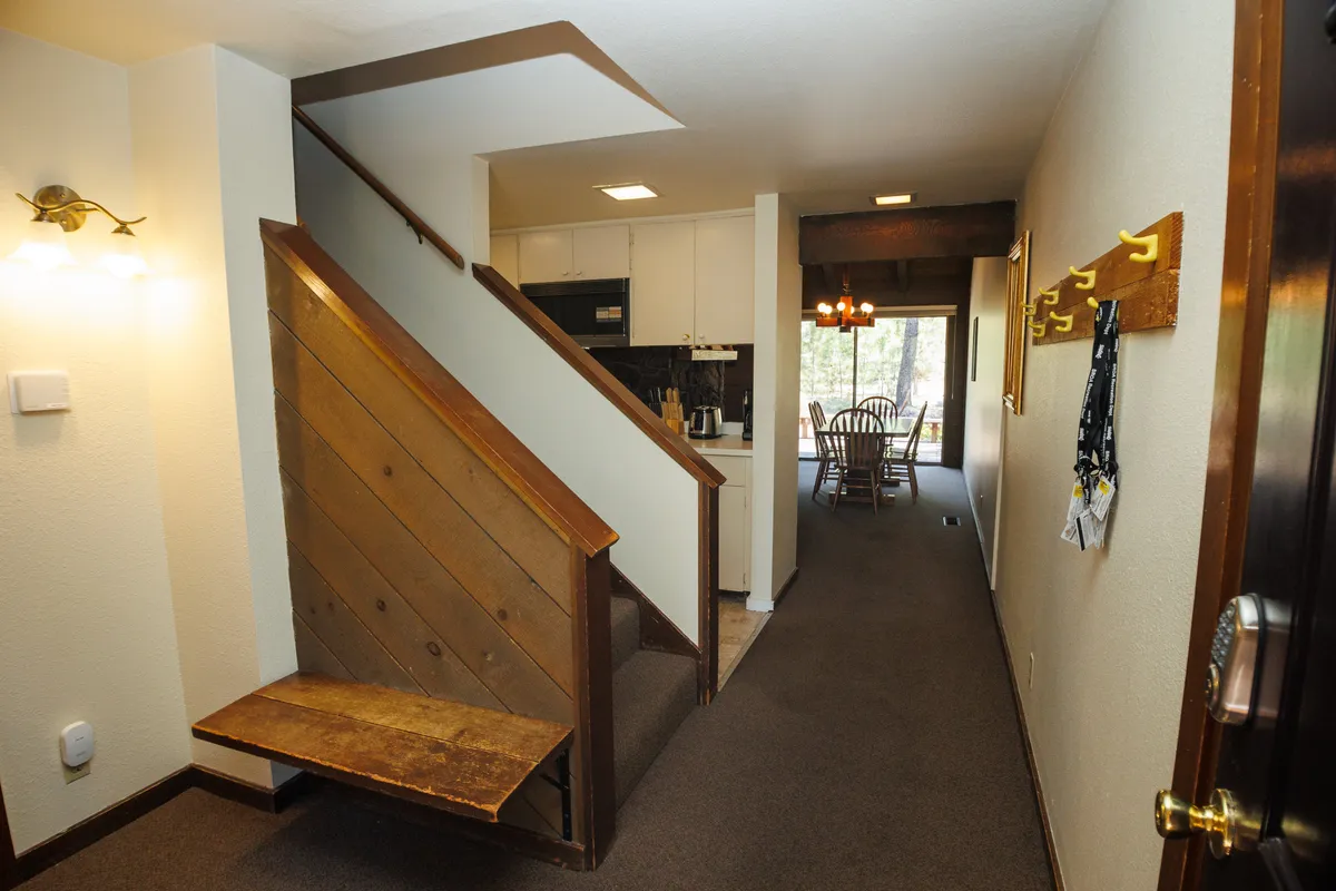 This image features a staircase leading to the entryway, a dining area visible in the background, wall hooks for hanging gear, and a bench for sit-down convenience. The space is open and inviting with plenty of natural light, suggesting easy movement between indoors and outdoors.